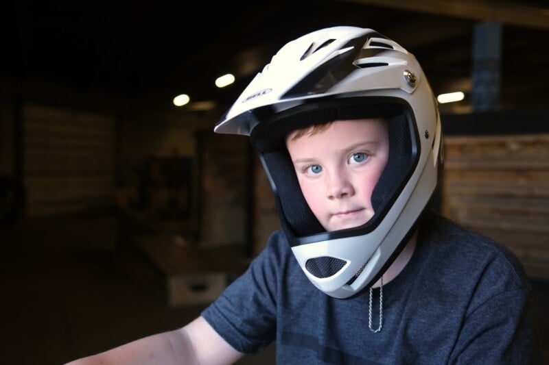 A young person wearing a white full-face helmet is looking at the camera. They are indoors in a dimly lit space with wooden structures and equipment in the background. They are wearing a dark gray shirt.