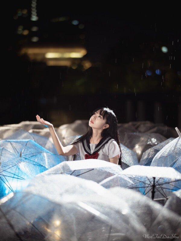 A young woman in a sailor-style school uniform stands amidst a sea of transparent umbrellas at night. She looks upwards with her right hand extended, as if feeling for rain. The background is dark with out-of-focus city lights.