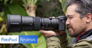 A person with gray hair and a beard is looking through the viewfinder of a camera with a large Fujinon lens. The setting appears to be outdoors with green foliage in the background. The image includes the text "PetaPixel Reviews" in the bottom left corner.