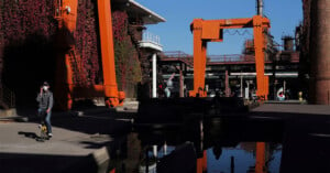 A person wearing a face mask rides a scooter next to a large industrial area with orange cranes and structures. The background features buildings covered in red ivy and a clear blue sky, reflected in a water body below.