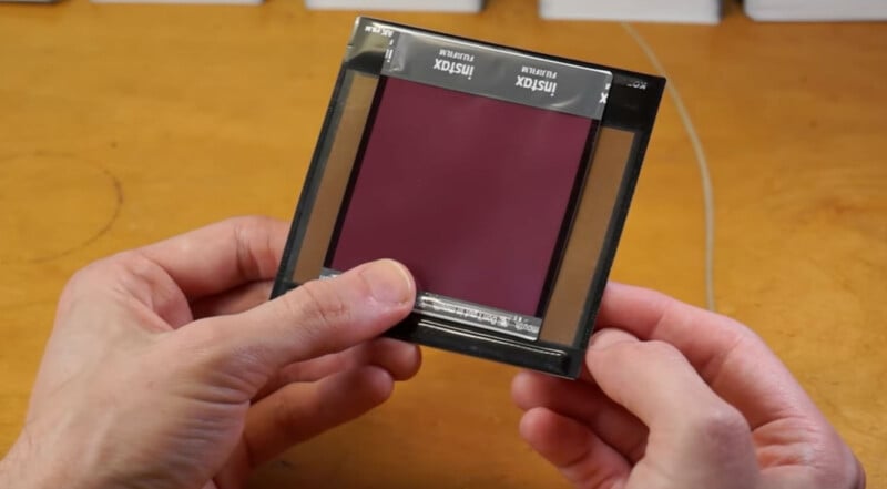 Close-up of two hands holding a dark red film negative slide with a black border. The slide is labeled "INISTIX" on the top edge, and is held over a wooden surface.