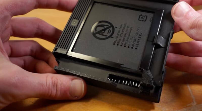 Close-up of a person's hands holding a partially disassembled battery casing. The battery is labeled with various text and symbols. The casing appears to be 3D-printed, as indicated by the visible layers and textured surface. The battery contacts and a spring are visible.