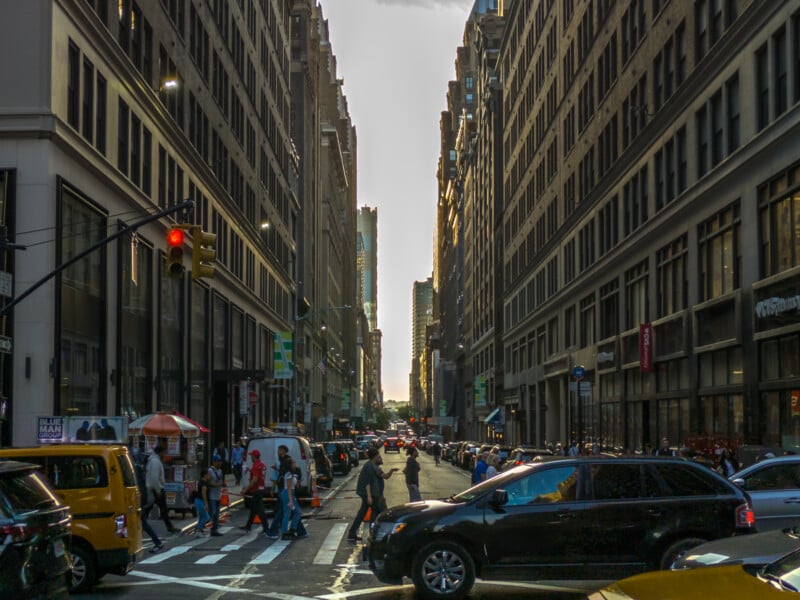 A bustling city street during late afternoon, with tall buildings on both sides. People are walking across the intersection, and various vehicles, including yellow taxis, fill the road. The sky is visible in the distance, casting a soft light over the scene.