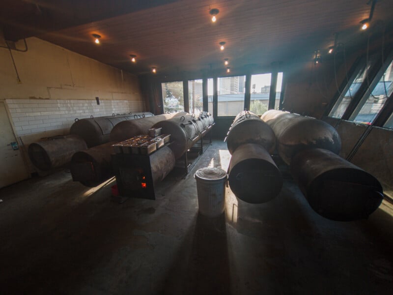 A dimly lit room with several large cylindrical metal smokers positioned in rows along the floor and by the wall. The room has a tiled wall on one side and multiple windows on the other, letting in some natural light. There is a bucket in the foreground.