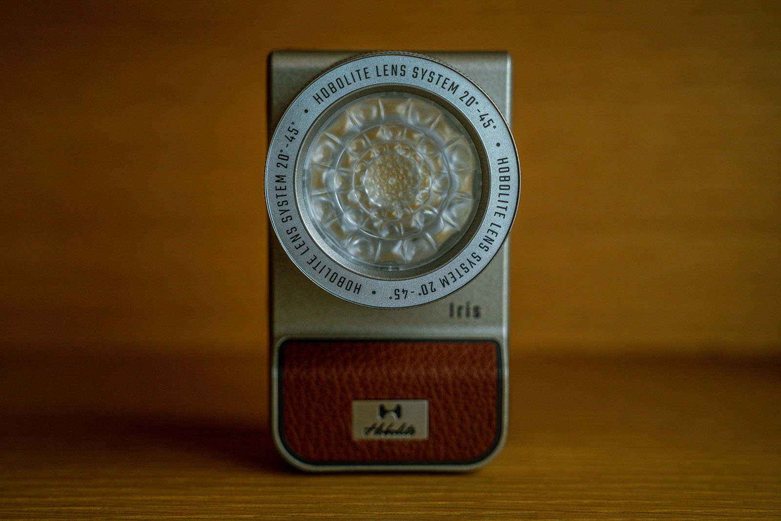 A Hobolite Iris lens system is displayed against a wooden background. The lens has a detailed, intricate design and is encased in a metallic rectangular frame with a brown textured base and the brand name "Hobolite" embossed on it.