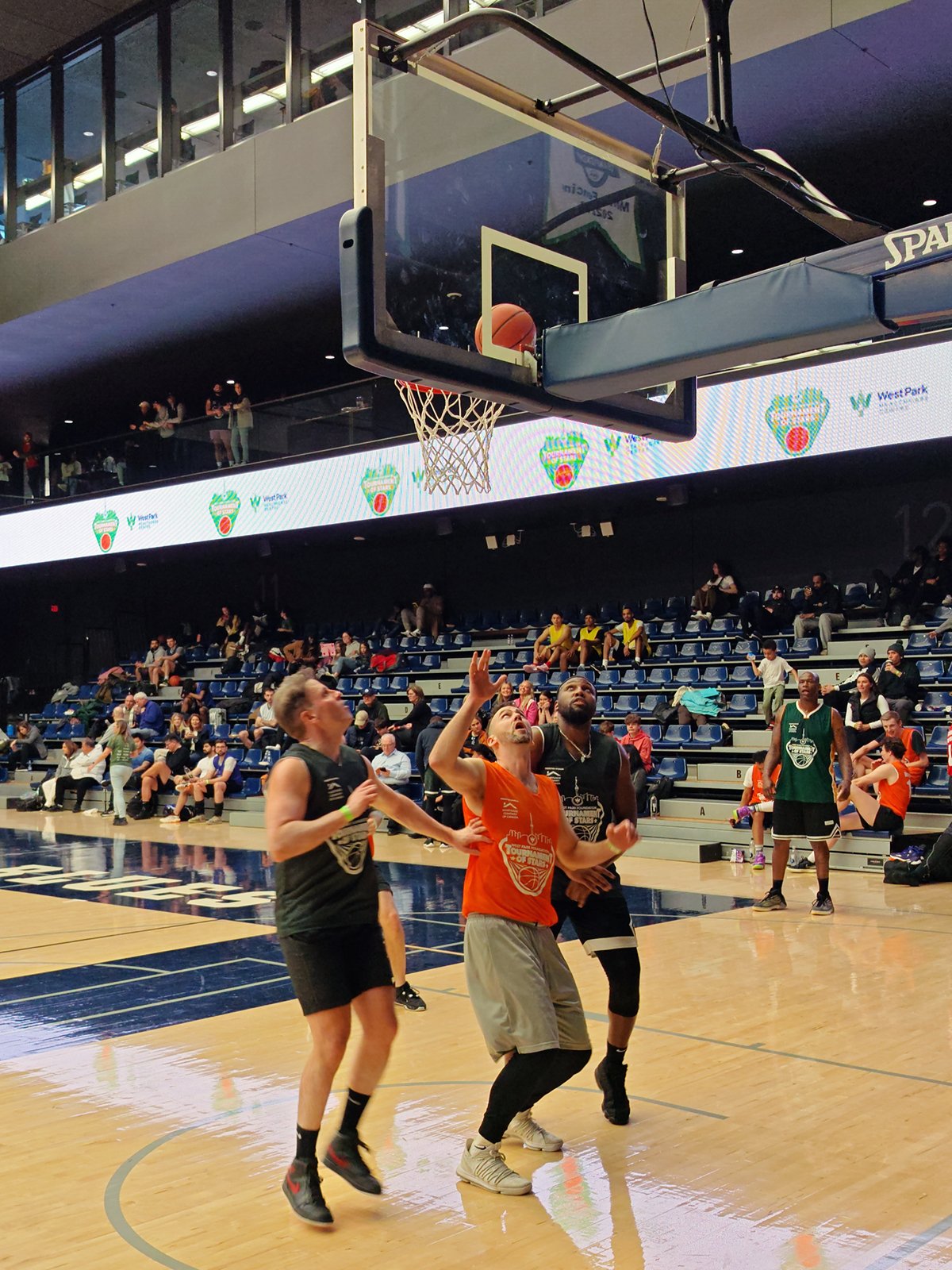 Three men playing basketball in an indoor court focus on a ball approaching the rim of the basket, with spectators in the background.