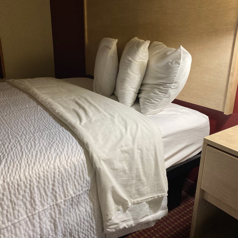 A neatly made bed with white sheets and four propped up pillows is in a dimly lit room with a bedside table. The headboard and wall behind the bed are a muted beige color, and the room carpet has a patterned, red design.