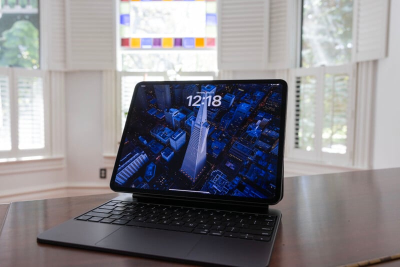 A laptop is open on a desk in a bright room with white shutters and stained glass windows, displaying an aerial night view of a city on its screen.