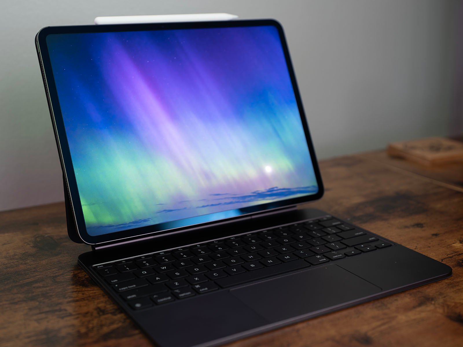 A tablet with a keyboard attached is placed on a wooden surface. The tablet screen displays an image of colorful aurora lights in shades of blue, green, and purple. The tablet is positioned at an angle, with a light gray wall in the background.