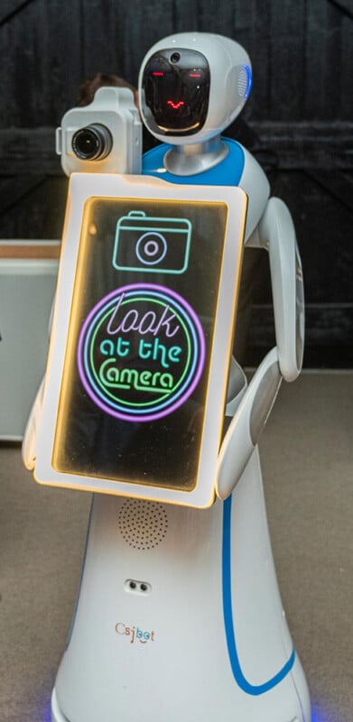 A humanoid robot with a screen on its chest displaying the message "Look at the Camera" holds a camera. The robot has a white and blue body with an expressive digital face and stands indoors against a dark background.