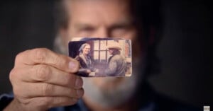 A man holds an old, slightly worn photograph up to the camera. The photo shows a younger man and woman smiling and interacting outdoors. The background is blurred, focusing attention on the man’s hand and the photograph he is holding.