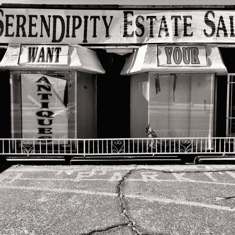 A black-and-white photo of a storefront titled "Serendipity Estate Sales." The left window displays the word "WANT" above a sign that reads "ANTIQUE." The right window displays the word "YOUR." The sidewalk in front appears weathered with visible cracks.