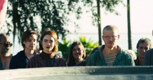 A group of people stand outdoors, facing the camera with neutral expressions. The setting is sunny and there are trees in the background. The group is diverse, including individuals with different hair colors and styles, with some wearing glasses.