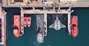 Aerial view of a dock with five vessels moored. Two large red boats are docked at either end, with three smaller boats between them. The central boat is a dark color, adjacent to a green vessel with visible solar panels. The dock is surrounded by water.