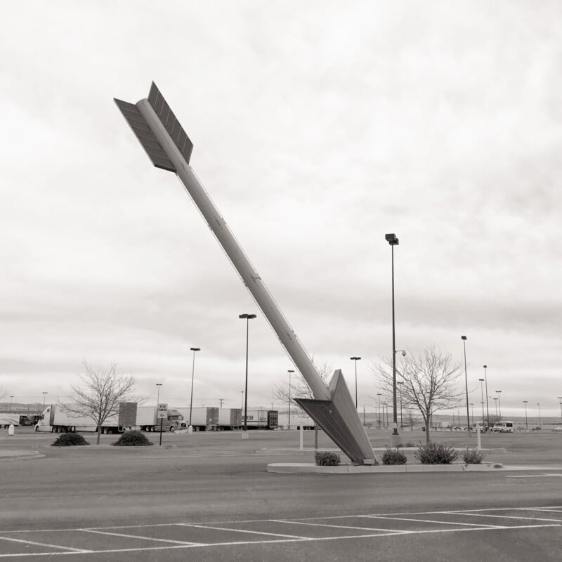 A giant arrow sculpture is embedded diagonally into the concrete in a mostly empty parking lot. The sky is cloudy, and there are a few barren trees and streetlights in the background.