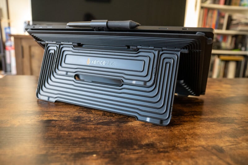 A stack of XenceLabs drawing tablets is neatly arranged on a wooden table. A stylus rests on top of the stack. In the background, shelves filled with books and a television are visible.