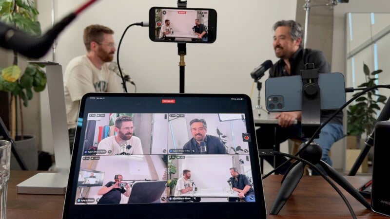 Two men are seated at a table, each speaking into a microphone. In the foreground, a tablet and a smartphone display multiple camera angles of the conversation. The setup includes various recording equipment and a few plants in the background.