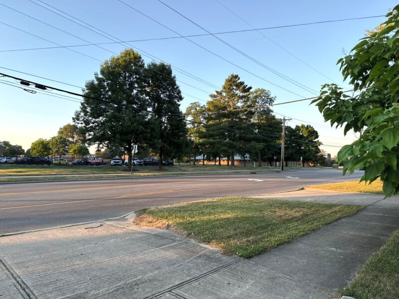 A quiet suburban street with a grassy verge and sidewalk in the foreground. Large trees line the road, casting shadows in the evening light. Power lines cross the sky, and in the distance, there are parked cars and a fenced area.