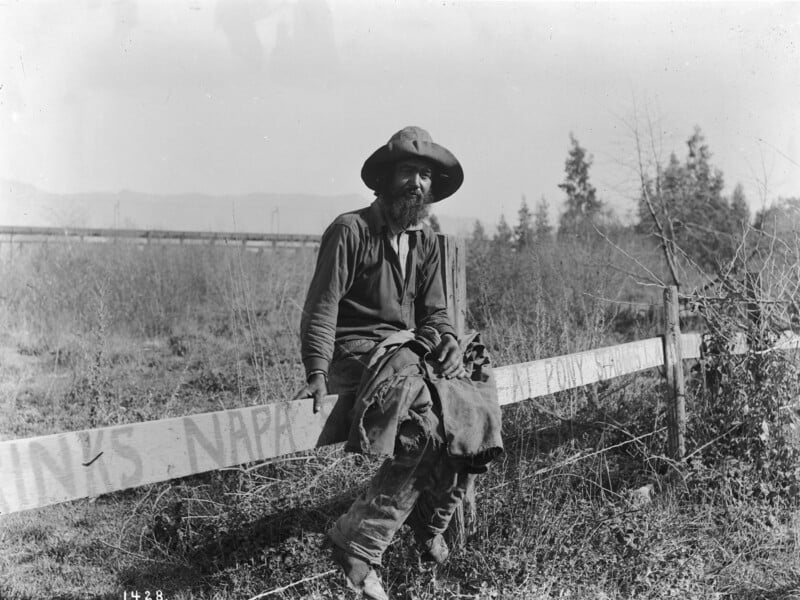 A man with a beard and a wide-brimmed hat sits casually on a wooden fence in a rural setting. He rests his arms on his lap, and the fence has text partially visible. The background features trees, grass, and a hazy sky.