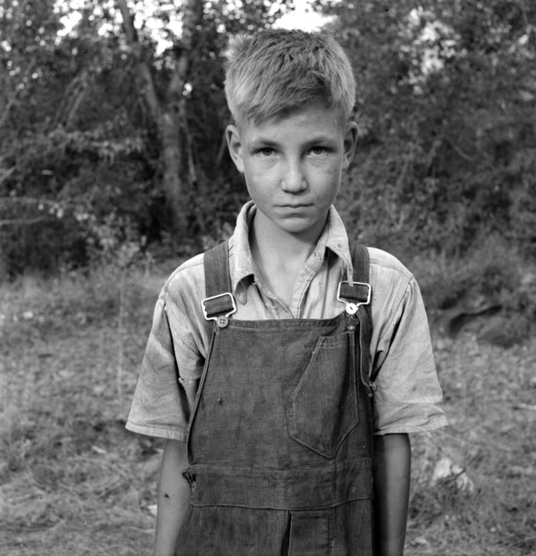 A young boy with short blond hair stands in front of a wooded area. He is wearing a light-colored shirt and overalls, with a serious expression on his face. The background consists of blurred trees and foliage.