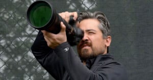 A man with graying hair and a beard intently looks through a large zoom lens camera, aiming it upward. He wears a black jacket, and the background features a green mesh fence. The camera is pixelated, obscuring its make and model.