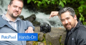 Two men are standing outdoors near a flowing stream, and one of them is pointing at a camera placed on a stone surface. The overlay text reads "PetaPixel Hands-On." Both men are wearing jackets, and greenery is visible in the background.