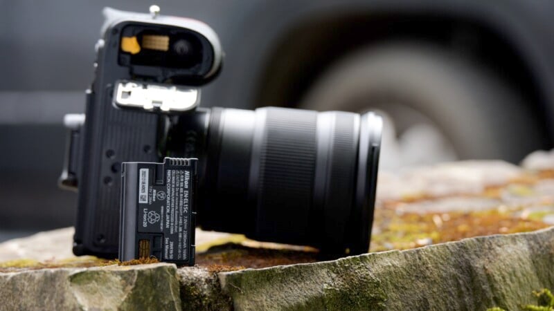 A black DSLR camera placed on a mossy, rough-textured rock with its memory card slot and battery compartment open. The battery is partially ejected and rests against the camera. The background is blurred, focusing attention on the camera.