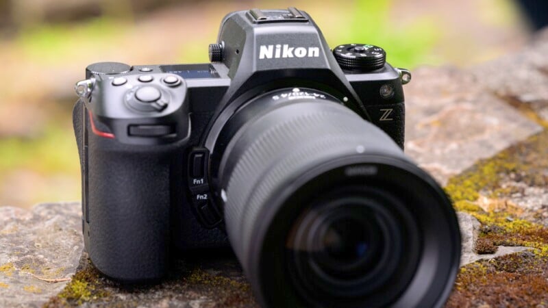 A Nikon Z-series mirrorless camera rests on a mossy rock surface. The camera is black with a textured grip and features multiple control buttons and dials. The lens is prominently extended, indicating the camera is ready for shooting.