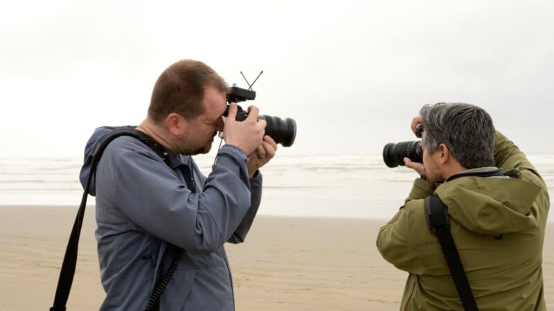 Two people stand on a beach, facing each other and taking photos with their cameras. The person on the left wears a blue jacket, while the person on the right wears a green jacket and glasses. The background features a sandy beach and a cloudy sky with ocean waves.