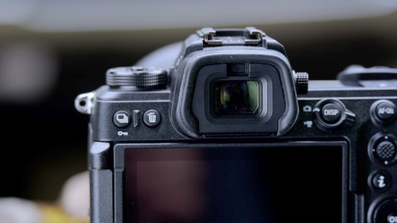 A close-up view of the back of a black digital camera, focusing on its viewfinder and LCD screen. Various control buttons and dials are visible around the viewfinder and screen. The background is blurred, putting emphasis on the camera details.