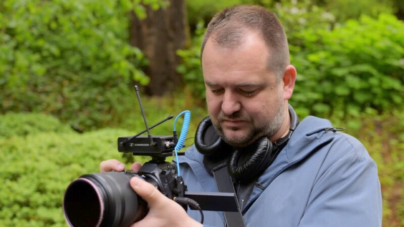 A man with headphones around his neck focuses on adjusting a camera with a microphone attached. He is outdoors, surrounded by green foliage, and wears a blue jacket.