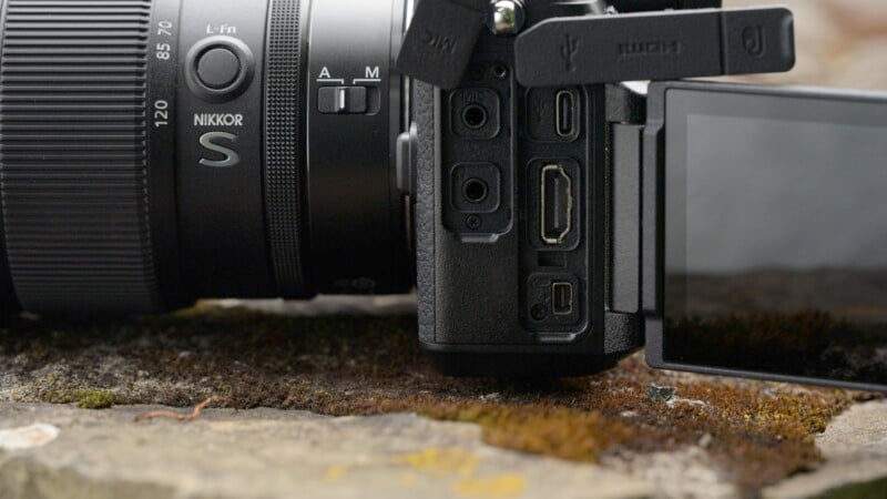 Close-up view of a digital camera with an external screen extended, exposing various ports including HDMI, USB, and microphone jacks. The camera is placed on a mossy rock surface, with a Nikkor S lens visible.