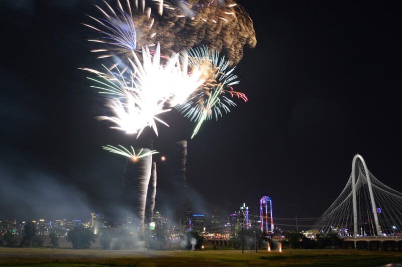 A nighttime cityscape featuring vibrant fireworks illuminating the sky. The scene includes a well-lit modern suspension bridge on the right, colorful illuminated buildings, and a green field in the foreground partially obscured by smoke.