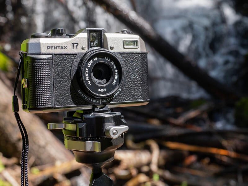 A vintage Pentax 17 film camera mounted on a tripod is positioned in front of a blurred waterfall in a forested area. The camera has a sleek, classic design with black and silver details.