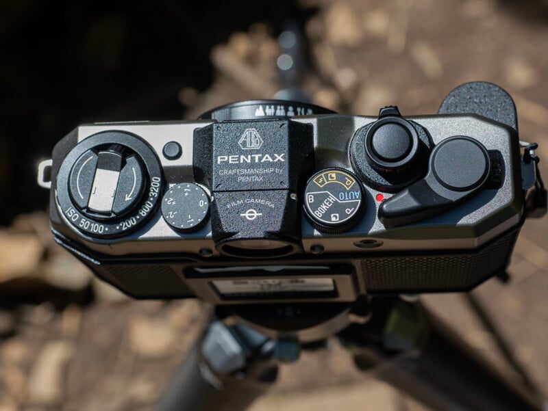 Top view of a Pentax camera on a tripod. Visible controls include the mode dial, ISO dial ranging from 100 to 3200, shutter release button, and various other buttons and switches for camera settings. The branding "Asahi Pentax" is prominently displayed.