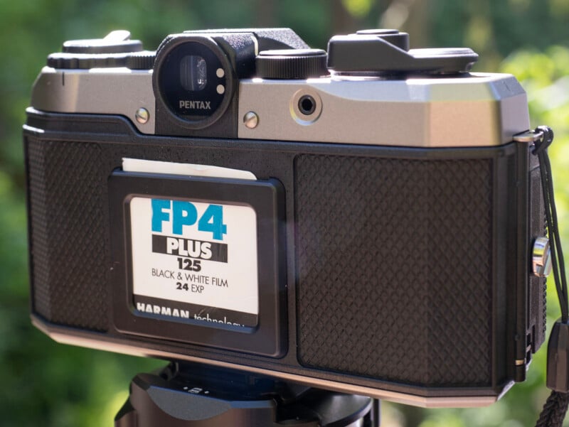 A close-up shot of the back of a vintage Pentax camera mounted on a tripod. The camera has an Ilford FP4 Plus 125 black and white film case inserted in the film slot. The background is blurred with hints of greenery.