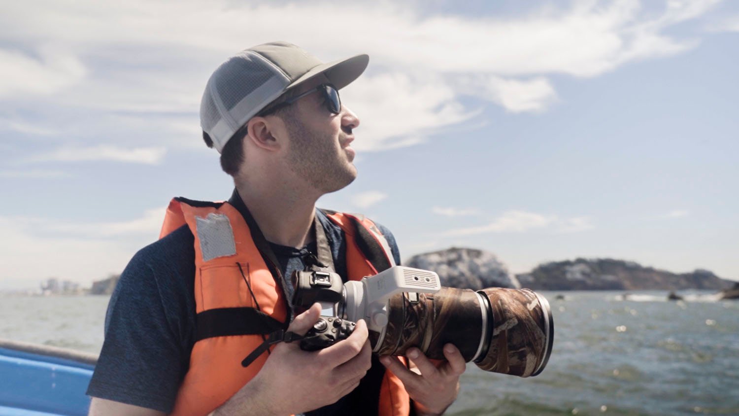 A man in a life jacket and cap holds a camera, looking out to sea from a boat, with a hilly island visible in the distance under a bright sky.