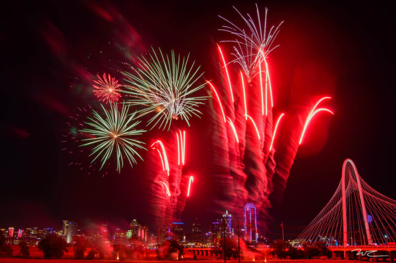 A night sky vibrant with fireworks of various colors, including red and green, illuminating a cityscape. A bridge is visible on the right side, with the city’s skyline in the background. The fireworks create dynamic trails of light against the dark backdrop.