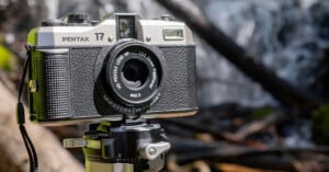 A Pentax 17 film camera mounted on a tripod stands outdoors with a blurred waterfall in the background. The camera has a classic compact design with a black and silver body, and a 1:3.5 45mm Pentax lens. The scene is set in a natural environment with logs and rocks.