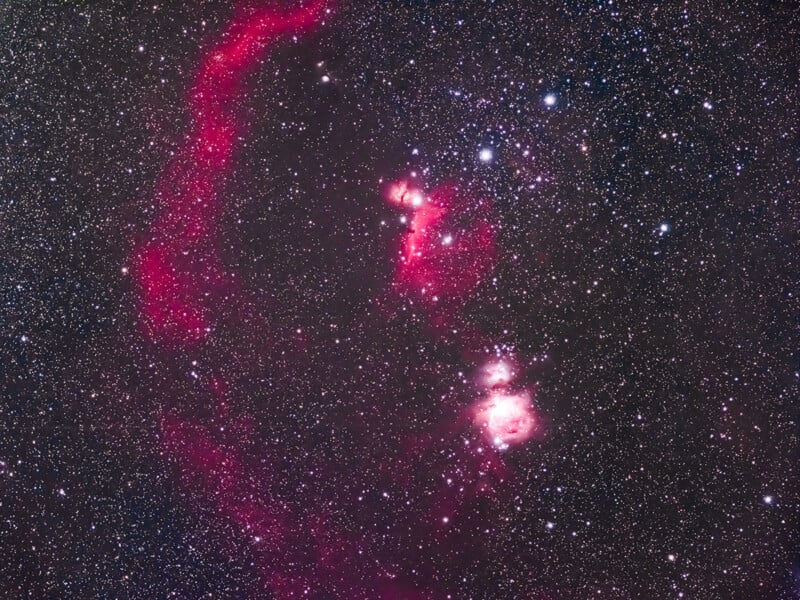 A vibrant, star-filled night sky featuring the Orion Nebula. The image displays countless stars scattered across the dark expanse, with noticeable red and pink hues of cosmic gases and dust forming intricate patterns, creating a stunning celestial view.