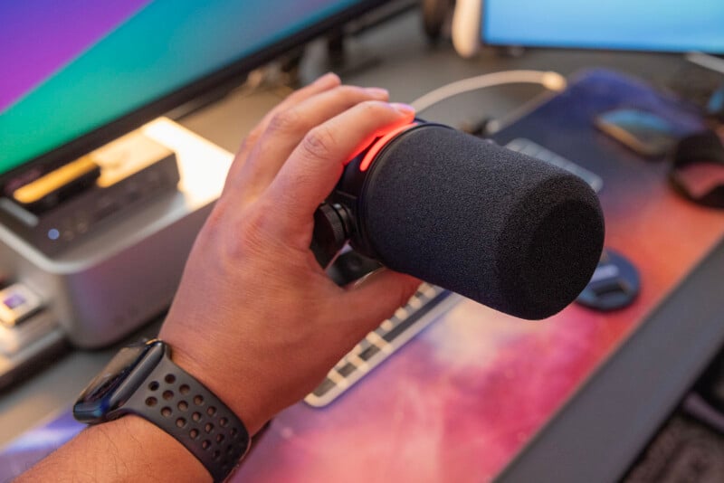 A person holding a black microphone with a foam cover near a computer setup with a colorful screen in the background.