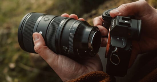 A person is holding a Sony camera in their right hand and a detached Sigma lens in their left hand. The background appears to be an outdoor setting with greenery.