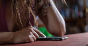 A person with long hair leans on a table, their face not visible. They are using a smartphone with their right hand, wearing bracelets on their left wrist. The background is blurred, showing indistinct shapes and colors.