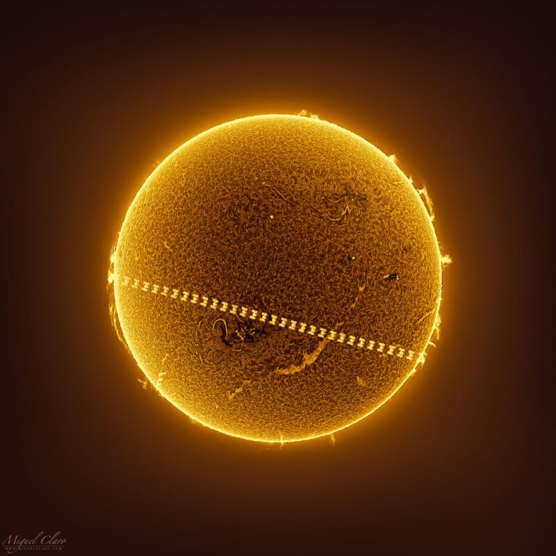 An image of the Sun with a bright, golden-yellow appearance against a dark background. The surface is detailed with visible texture and solar flares. A diagonal, dotted line runs across the Sun, representing the transit of the International Space Station (ISS).