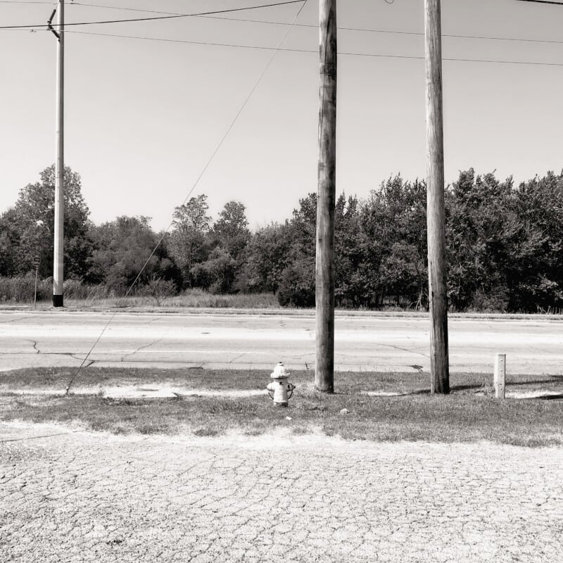 A black and white image of a rural roadside scene. Two tall utility poles stand alongside a small fire hydrant on a grassy patch. A paved road is visible in the background, with trees and bushes lining the horizon under an open sky.