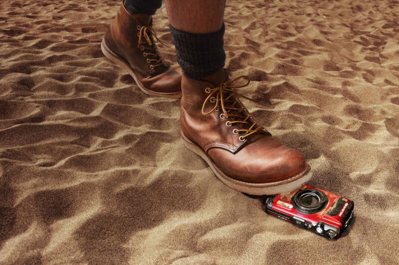 A person wearing leather boots stands on sandy ground, one boot pressing down on a camera partially buried in sand.