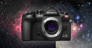 A black Olympus OM-D E-M1 Mark III camera with a textured grip is displayed against a vibrant, colorful background of outer space with stars and nebulae. The camera is shown without a lens, revealing the sensor inside.