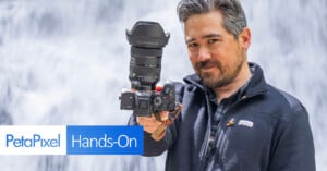 A man holding a professional camera with a large lens is standing in front of a waterfall. He is wearing a dark jacket, and there is a "PetaPixel Hands-On" graphic at the bottom left corner of the image.