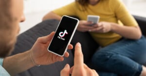 A man and a woman sitting on a sofa, each using their smartphones. the man's phone, held in his hand, displays the tiktok logo on its screen. focus on the man's phone screen.