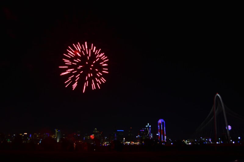 A nighttime scene with a vibrant red firework exploding in the sky. Below, the illuminated skyline of a city is visible, including a distinctive bridge with a tall arch. The cityscape features various brightly-lit buildings and structures.